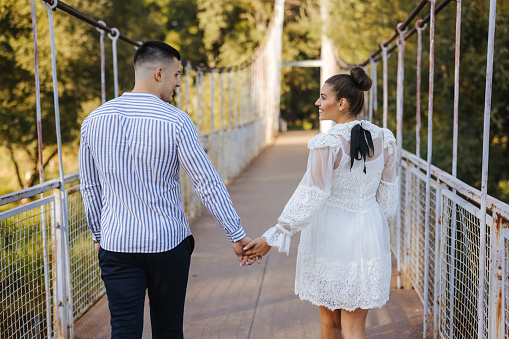 Back view of young romantic couple holding hands and walking along footbridge on sunny day