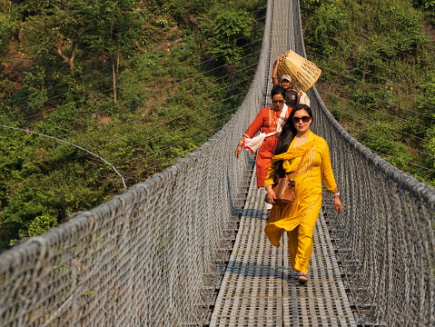 Chitwan district, Nepal - April 25, 2022 : Nepali women dressed in traditional colorful clothes are walking on a suspension bridge. Behind them, a man is currying a basket. Suspension bridges are common in Nepal. People use them to cross the rivers on foot or with their motorcycles.