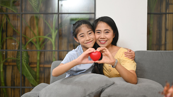 Little girl and mother holding heart shape and smiling at camera. Life insurance, love and support in family relationships concept.
