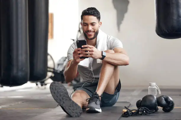 Photo of One fit young hispanic man listening to music with earphones from a cellphone while taking a break from exercise in a gym. Happy guy texting, making video call and using fitness apps online while browsing social media and watching workout tutorials