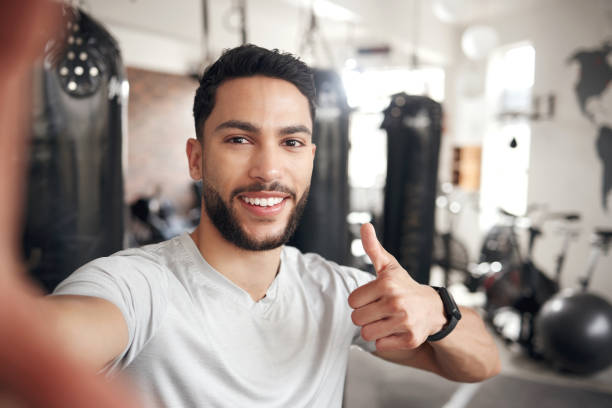 istock Portrait of one confident young hispanic man taking selfies and gesturing thumbs up while exercising in a gym. Happy mixed race instructor looking motivated and ready for a good training workout in a fitness centre 1399520067