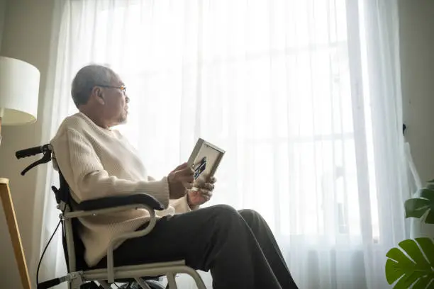 Photo of Asian senior male sit alone on wheelchair and looks family photographs. Elderly older mature grandfather feel lonely and sad, missing his relative and wife while stay in nursing home after retirement.