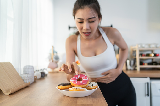 Asian beautiful young girl hungry and want to eat donut in kitchen. Attractive woman wear sport cloth feeling famished after exercies and pick up junk food unhealthy to eat, Food and health concept.
