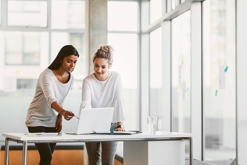 Young Indian ethnicity business woman and colleague standing in front of desk and working on laptop in modern business office