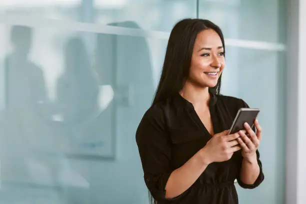 Photo of Mixed race multiracial Asian Hispanic Chinese woman texting on phone while looking off to the side in modern business office