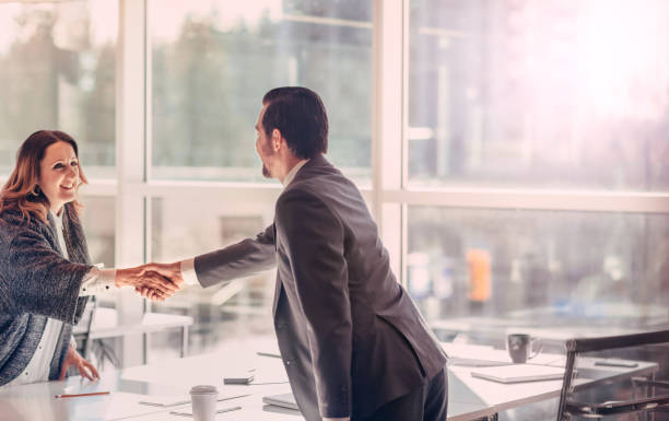 Man and woman standing shaking hands across a table in agreement job interview success inside a modern business office stock photo