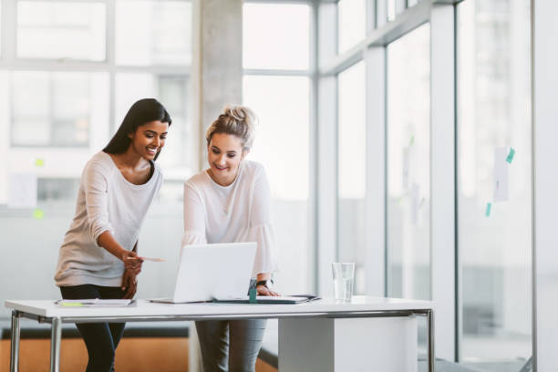 giovane donna d'affari di etnia indiana e collega in piedi davanti alla scrivania e lavorando sul computer portatile nel moderno ufficio aziendale - brainstorming team learning business foto e immagini stock