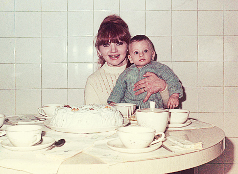 A vintage and grainy image of a cute baby boy with a smiling mom in the dining room in front of a big cake and teacups on the table.