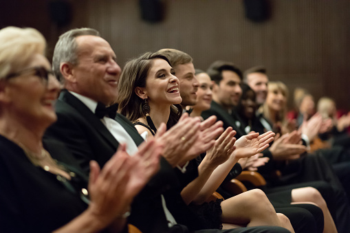 Side view of audience clapping hands in opera house. Men and women are watching theatrical performance. They are in elegant wear.