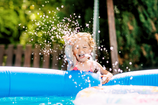 A cute baby girl splashes water in an inflatable pool in the garden and laughs. Summer, water, spray, children.