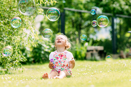A little girl blows soap bubbles in the park, sitting on the grass. The concept of a happy childhood.