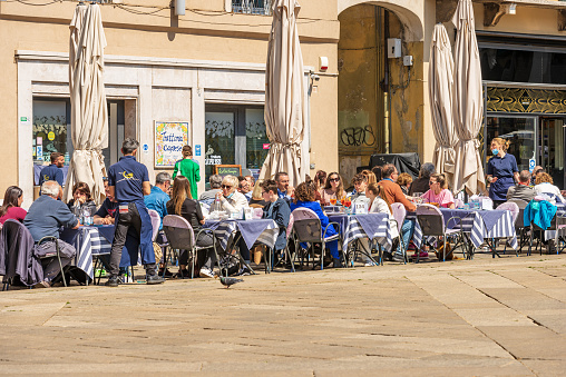 Brescia, Italy - April 17, 2022: Outdoor restaurant crowded with people on a sunny spring day in Brescia downtown, Loggia town square (Piazza della Loggia), Lombardy, Italy, Europe. This square in Brescia is famous for its ancient Renaissance-style building Loggia Palace (Palazzo della Loggia), 1492-1574, and for the tower with the astronomical clock, 1540-1550.
