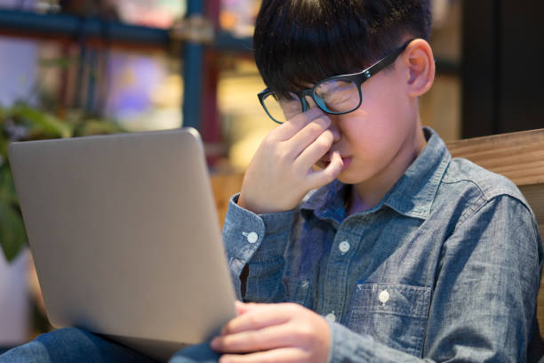 lindo niño asiático de escuela primaria con anteojos frotándose la nariz y los ojos mientras estudia la clase en línea con computadora portátil en la escuela - frotarse los ojos fotografías e imágenes de stock