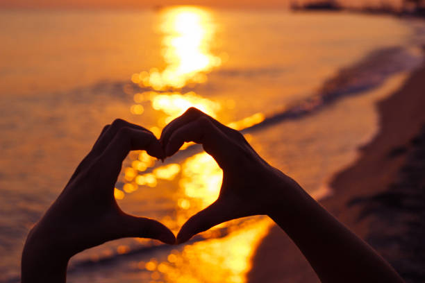 Hand holding heart shape against sky during sunset