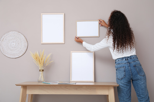 African American woman hanging empty frame on pale rose wall over table in room, back view. Mockup for design