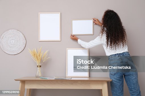 istock African American woman hanging empty frame on pale rose wall over table in room, back view. Mockup for design 1399559010