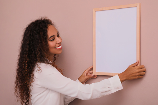 Happy African American woman hanging empty frame on pale rose wall. Mockup for design