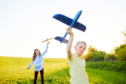 Running boy and girl holding two yellow and blue airplanes toy in the field during summer sunny day