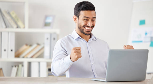 istock Young happy and excited mixed race businessman cheering in support while working on a laptop alone in an office at work. One hispanic male boss smiling while celebrating success and victory 1399562192