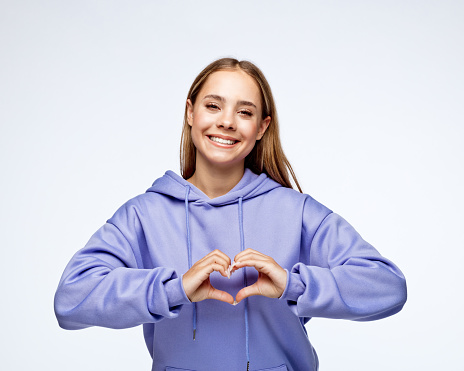 Portrait of smiling teenage girl gesturing heart shape against white background.
