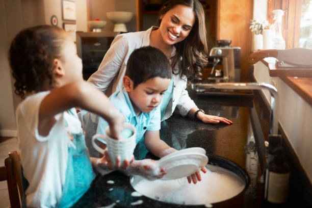 joven madre mestiza feliz lavando los platos con su hijo y su hija en la cocina de casa. hermano pequeño y hermana ayudando a su mamá con los platos. familia manteniendo su casa limpia - lavar los platos fotografías e imágenes de stock
