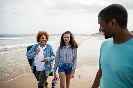 A multi-generation family walking near the water's edge at Beadnell beach, North East England. They are all smiling at each other while they walk and the grandmother is carrying a paddle board. The father is looking over his shoulder and smiling.
