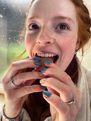 Stock photo showing close-up view of a young red-headed woman eating a chocolate flavoured macaron.