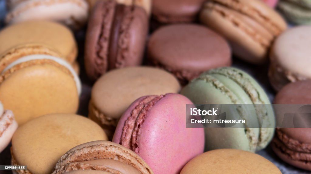 Full frame image of heap of multi coloured macarons filled with flavoured butter cream, vanilla, lemon, salted caramel, chocolate, strawberry and pistachio flavoured meringues, focus on foreground Stock photo showing close-up, elevated view a pile of multi coloured macarons on blue wood grain background. Vanilla, lemon, salted caramel, chocolate, strawberry and pistachio flavoured macarons ready for an afternoon tea party. Macaroon Stock Photo