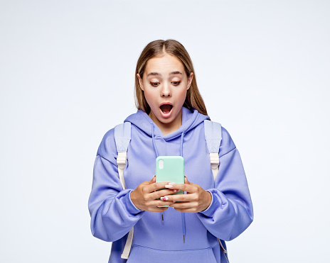 Portrait of surprised teenage girl with blond hair wearing lilac hoodie and backpack, holding smart phone, standing against white background.