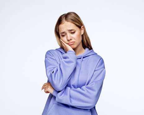 Depressed teenage girl wearing lilac hoodie with hand on chin standing against white background.