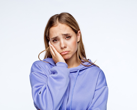 Depressed teenage girl wearing lilac hoodie with head in hand standing against white background.