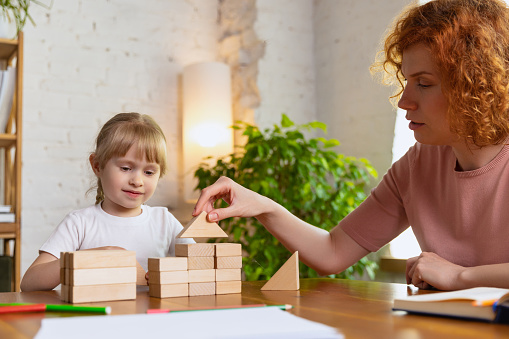 Develop fine motor skills and logical thinking. Smiling woman and cute little girl, mother and daughter playing with wooden toy cubes at home. Concept of family, support, studying, childhood, emotions