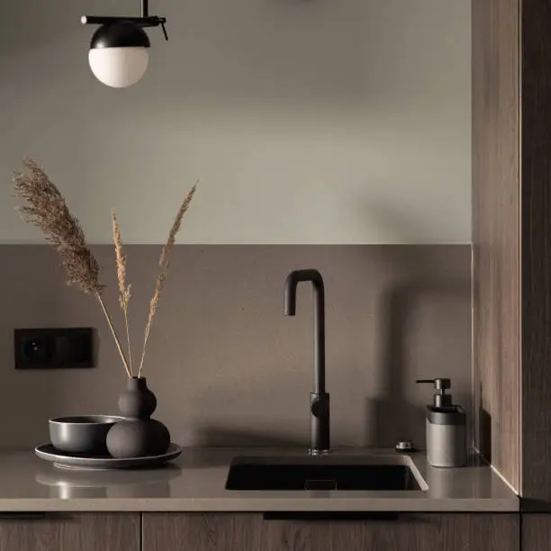 Close-up on stylish decorated kitchen countertop with black sink and tap, modern lamp and fancy vase, plate and bowl