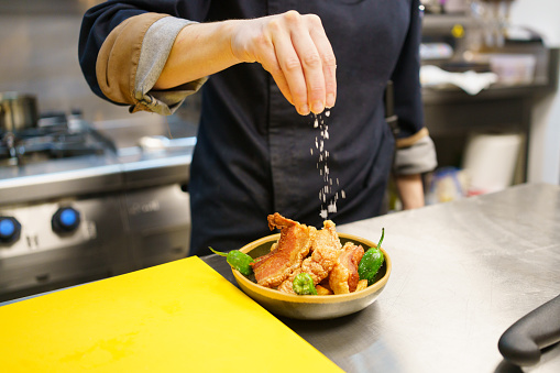 Unrecognizable cook sprinkling salt over a bowl of pork and hot peppers while working in the kitchen of a modern restaurant. Famous torreznos of Spanish cuisine