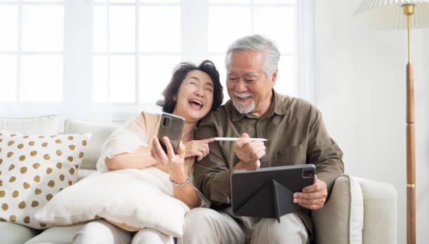 Asian senior couple in living room at home.Wife browsing online on smartphone showing something to her husband while husband is also using a tablet. stock photo