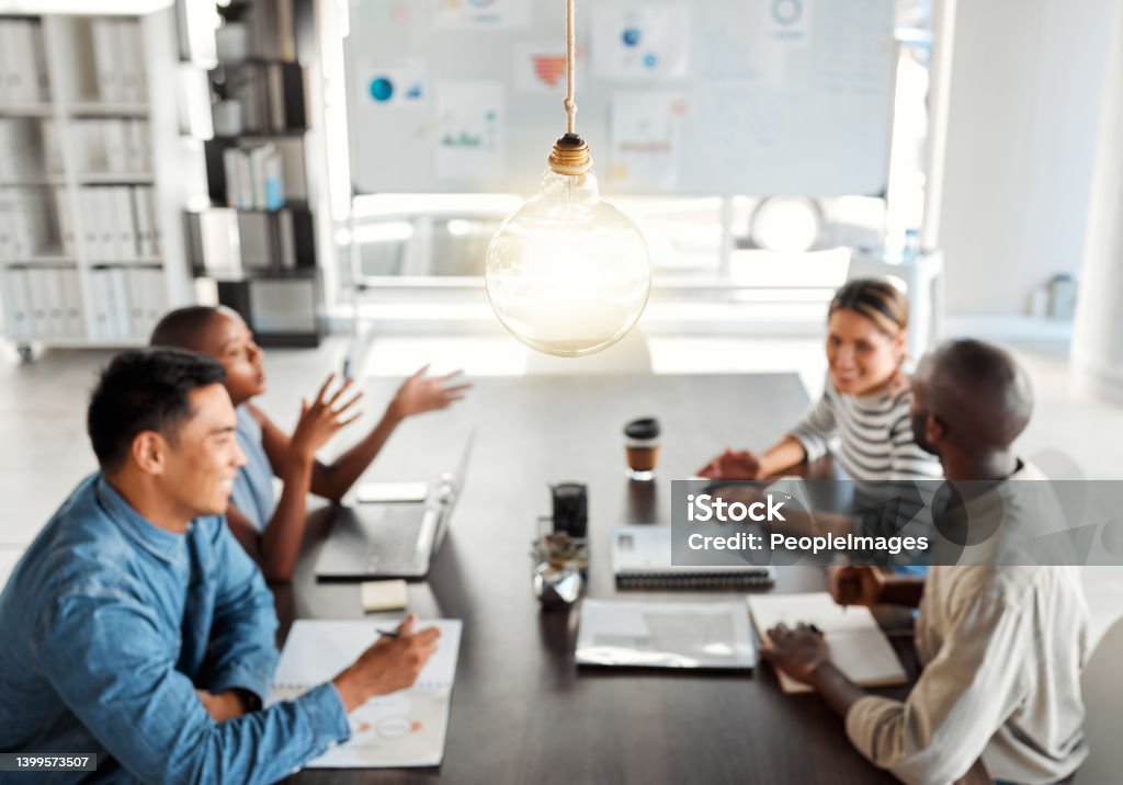 Bright lightbulb hanging from the roof while a group of businesspeople are having a meeting in an office at work. Bulb represents ideas, idea, creativity and inspiration. Colleagues planning together Office Stock Photo
