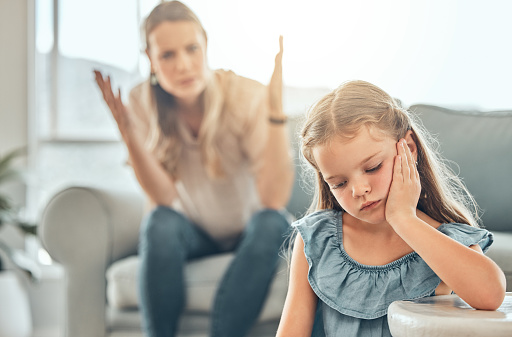 Sad little caucasian girl sitting in the living room and feeling depressed while her mother shouts and raises her hands behind her. Young cute daughter feeling upset after disappointing her parent at home