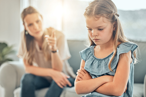 Closeup of an adorable little girl standing with arms crossed and looking upset while being scolded and reprimanded by her angry and disappointed mother at home. A woman punishing her young daughter