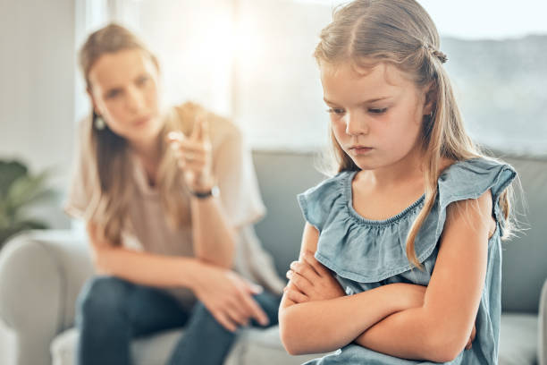 close-up de uma adorável garotinha de pé com os braços cruzados e parecendo chateada enquanto era repreendida e repreendida por sua mãe irritada e desapontada em casa. uma mulher punindo sua filha. - amuado - fotografias e filmes do acervo