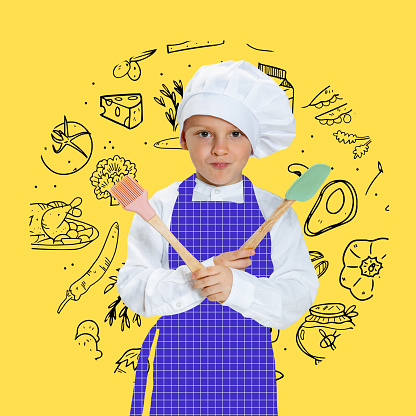 At cooking class. Art portrait of little happy boy in white cook uniform and huge chef's hat looking at camera isolated on yellow background with drawings. Childhood, studying, jobs, games, emotions.