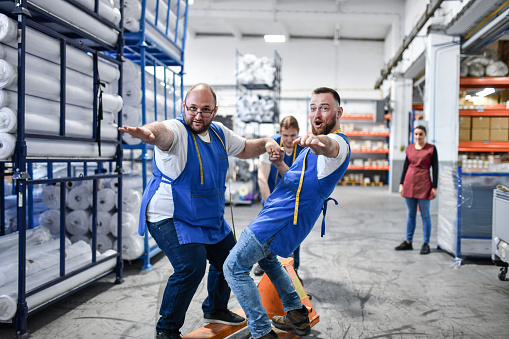 Two Male Warehouse Workers Balancing Themselves On Forklift Pushed By Colleague