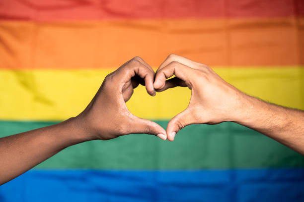 close up shot of hands making love or heart symbol in front of pride or rainbow flag - concept of gay couple love, solidarity and pride month celebration. close up shot of hands making love or heart symbol in front of pride or rainbow flag - concept of gay couple love, solidarity and pride month celebration peace demonstration stock pictures, royalty-free photos & images