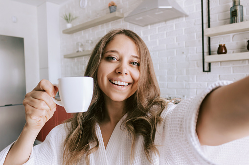 A young beautiful caucasian smiling blonde woman in a white robe with a white cup of coffee or tea takes a selfie in the morning on background of kitchen. Happy girl drinks hot beverage at home
