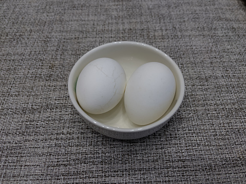 Two white boiled eggs in a small white bowl on a textured table. Selective focus on the egg and bowl.