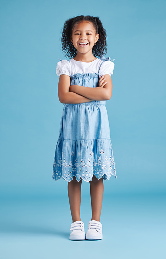 Retrato de una niña feliz de pie con los brazos cruzados sobre el fondo azul del estudio. Alegre niño mestizo con vestido vaquero casual y camiseta blanca photo