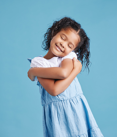 Adorable loveable little girl looking happy while hugging embracing herself. Cute kid with positive self-esteem against blue studio background