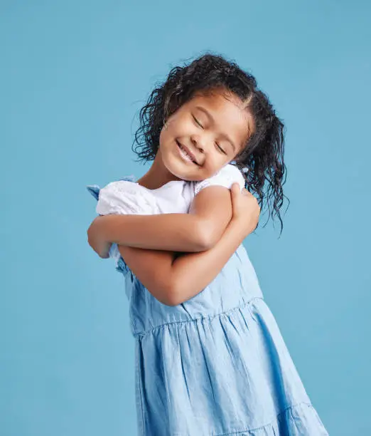 Photo of Adorable loveable little girl looking happy while hugging embracing herself. Cute kid with positive self-esteem against blue studio background