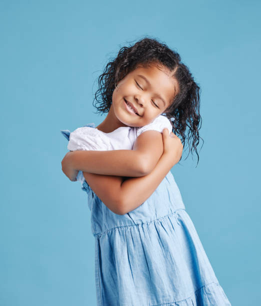 очаровательная милая маленькая девочка выглядит счастливой, обнимая себя. милый малыш с положительной самооценкой на синем студийном фоне - one kid only стоковые фото и изображения