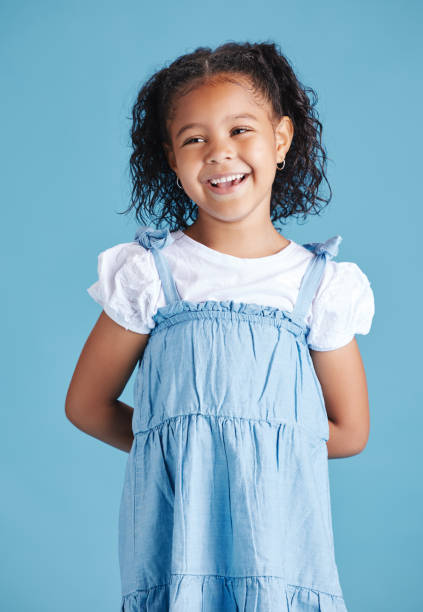 istock Happy smiling little girl standing with her hands behind her back against blue studio background. Cheerful mixed race kid in casual denim dress and white tshirt 1399611775