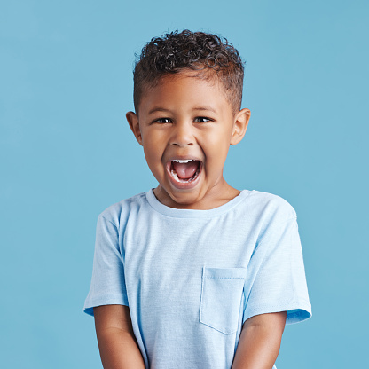 Close up of excited little boy looking happy and shouting while standing against a blue studio background. Cute preschooler wearing casual clothes